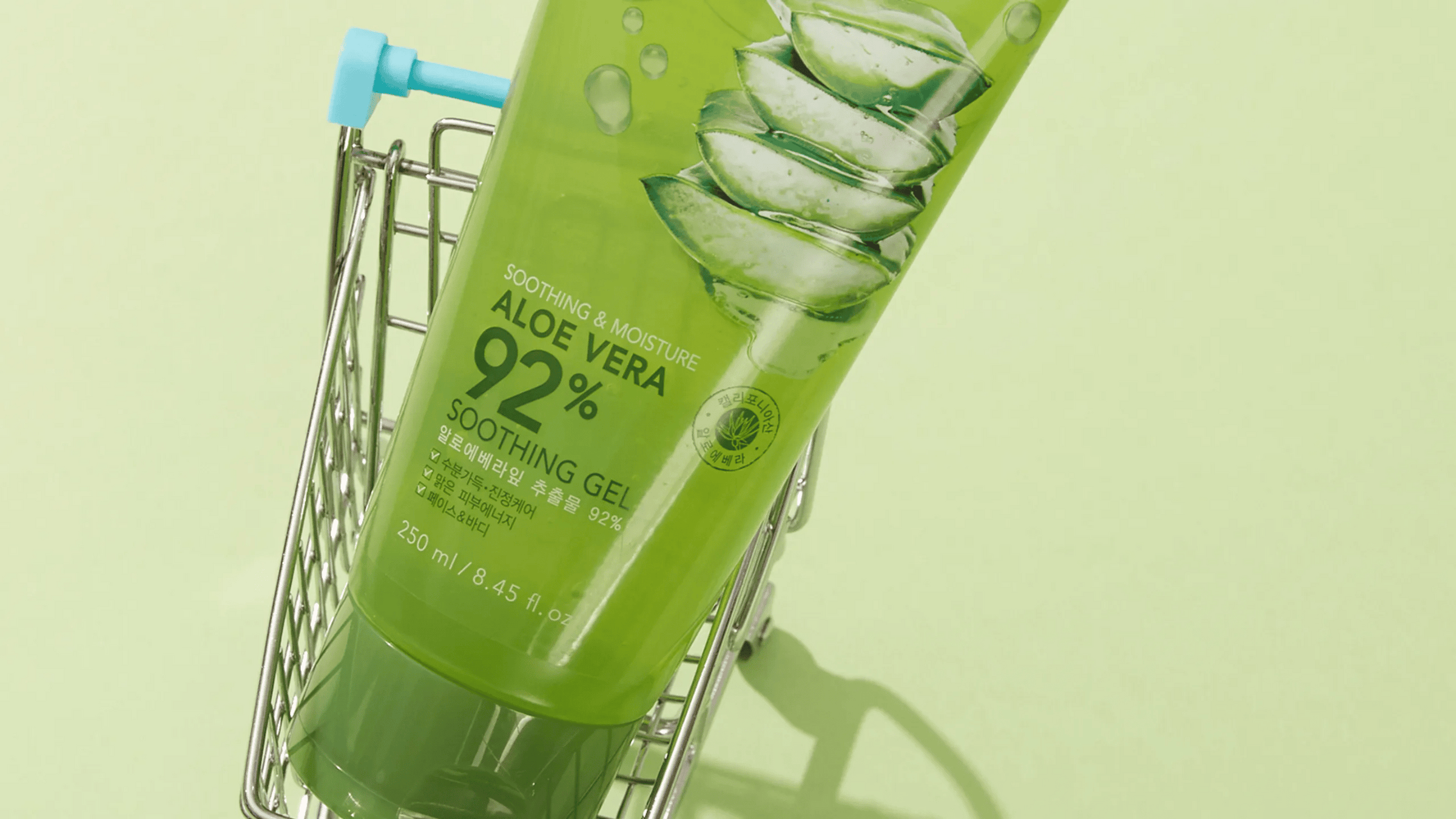 En grön tub med Nature Republic Aloe Vera 92% Soothing Gel ligger i en liten kundvagn mot en ljusgrön bakgrund. Förpackningen visar en bild av aloe vera och innehåller text om produktens lugnande och fuktgivande egenskaper.