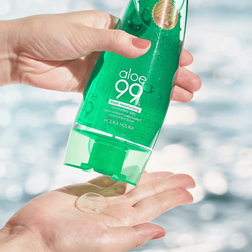 En hand håller en grön tub med Holika Holika Aloe 99% Soothing Gel, medan en annan hand fångar en klick gel som dispenseras. Bakgrunden är suddig och visar ett vattnigt, glittrande mönster.