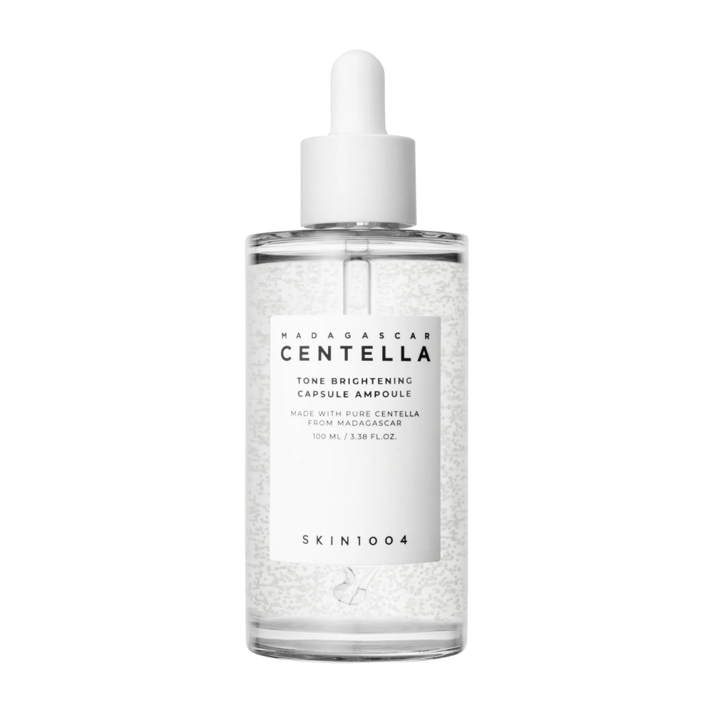 Bilden visar en flaska av "SKIN1004 Madagascar Centella Tone Brightening Capsule Ampoule". Det är en genomskinlig flaska med en vit droppapplikator. På etiketten står det att produkten innehåller rena centella-ingredienser från Madagaskar. Flaskans innehåll ser ut att ha små kapslar eller partiklar i sig, vilket kan vara en del av dess ljusnande hudvårdsegenskaper. Flaskan innehåller 100 ml / 3.38 fl.oz. av produkten.